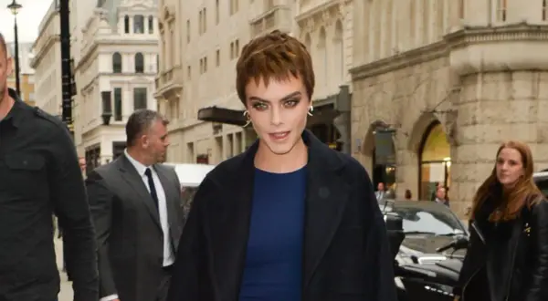 Cara Delevingne con un atrevido buzz cut largo, mirada intensa y abrigo oscuro, rodeada de seguridad en una calle urbana.