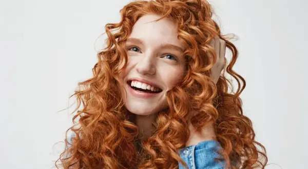 Mujer joven con cabello rojo y rizado, sonriendo y tocándose el pelo. Su melena merodo curly es espectacular.