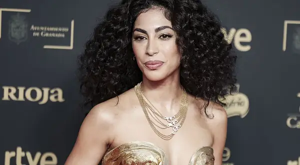 Mujer con cabello oscuro y rizos definidos, luciendo un vestido dorado y un collar de cadenas.