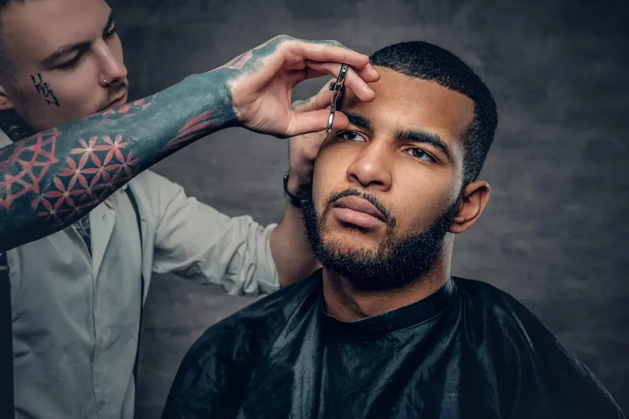 Barbero con tatuajes en el brazo recorta el **buzz cut corto** de un hombre con barba.