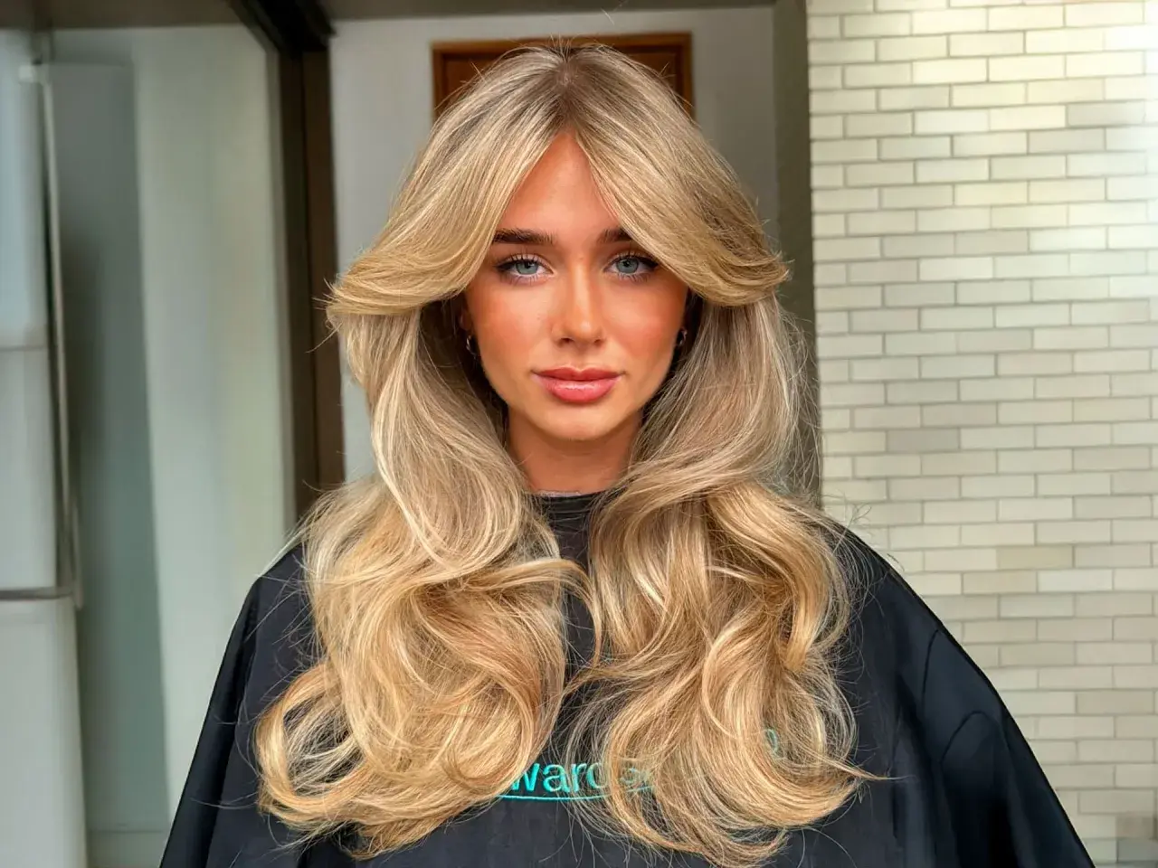 Mujer con cabello rubio largo y ondulado, con un flequillo mariposa que enmarca su rostro.