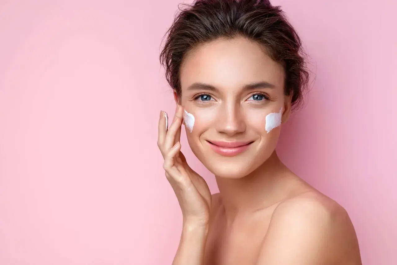 Mujer sonriente aplicándose crema en las mejillas, esencial para los cuidados de la piel en verano.