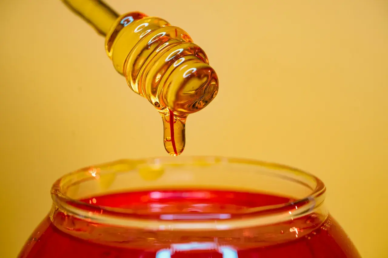 Miel dorada goteando de un agitador sobre un frasco rojo, ideal para un exfoliante facial casero.