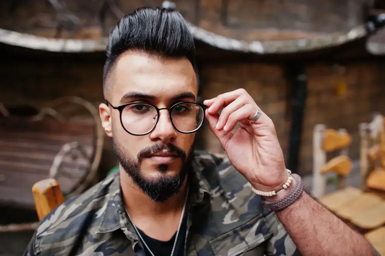 Hombre con barba y gafas, luciendo un moderno corte de pelo mohicano y chaqueta de camuflaje.