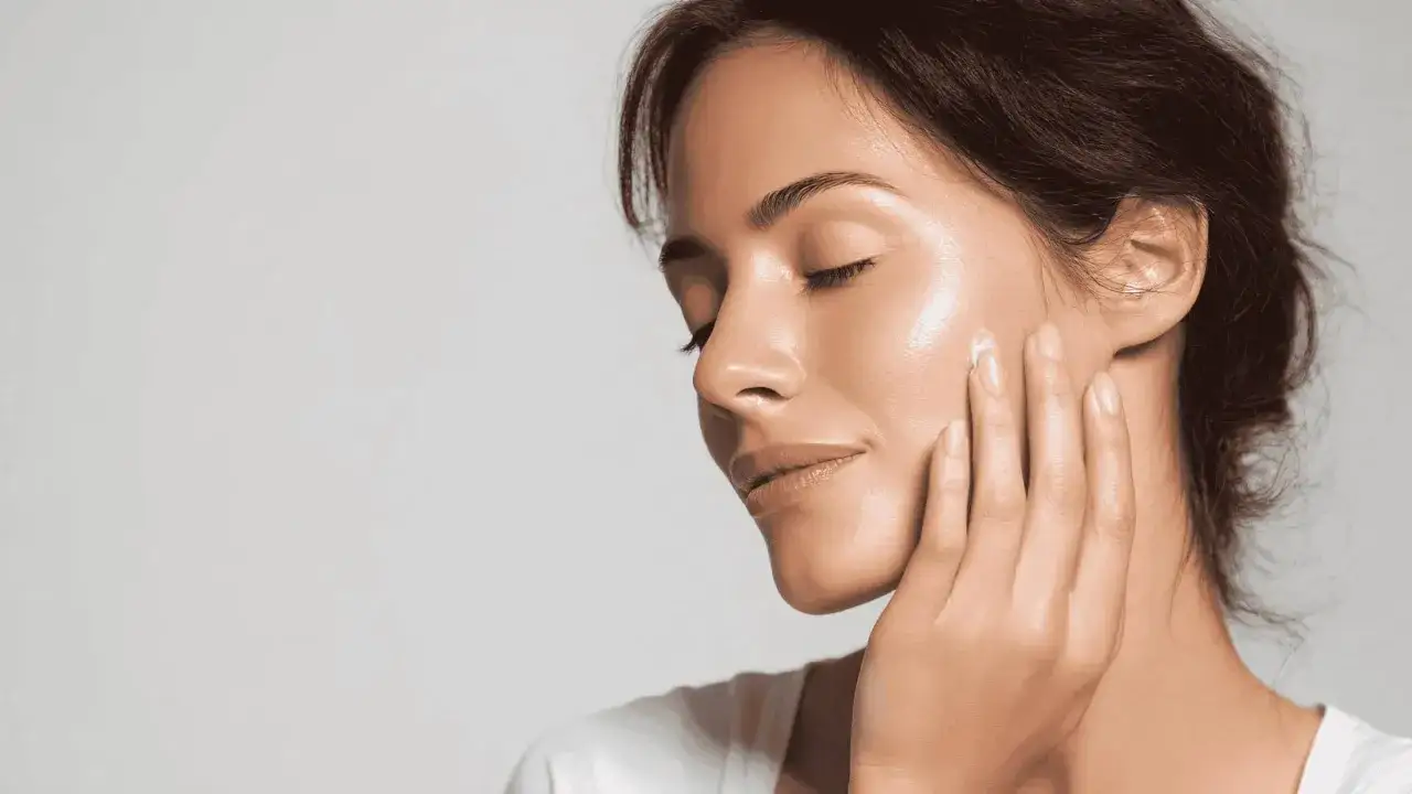 Mujer con ojos cerrados aplica crema en su rostro, buscando hidratar la piel de forma natural para un brillo saludable.