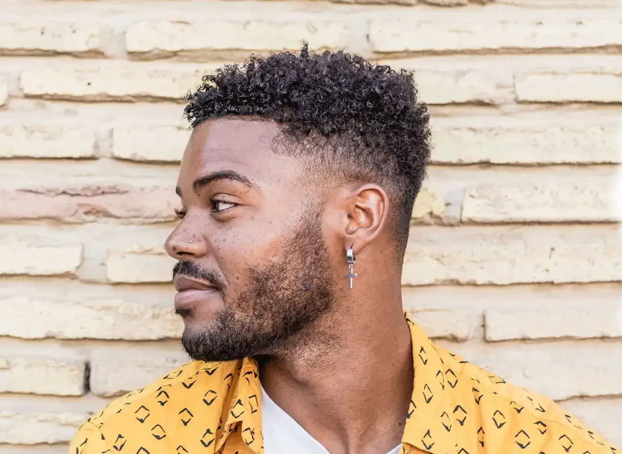 Hombre con un moderno corte taper fade, cabello rizado y barba cuidada, luciendo un pendiente de cruz.