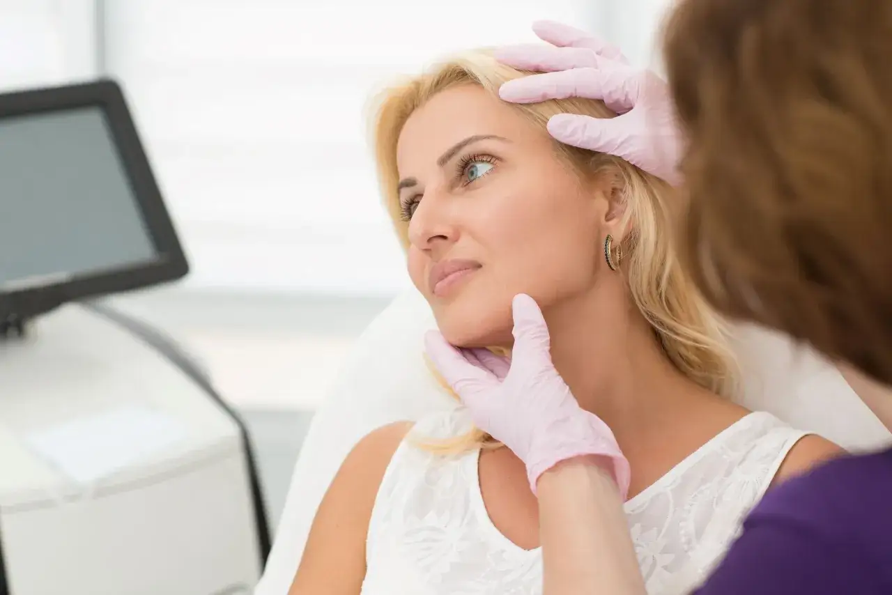 Manos enguantadas revisan el rostro de una mujer rubia, evaluando cómo alisar la piel de la cara para un tratamiento estético.