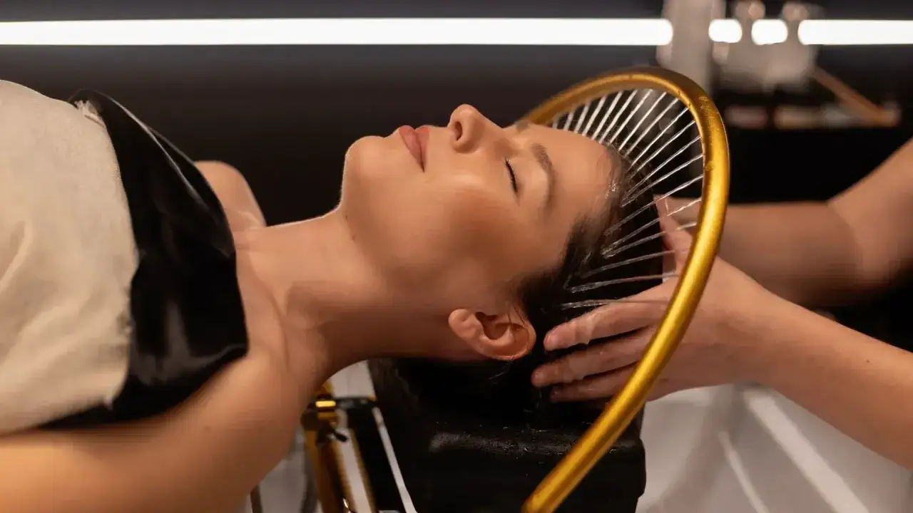 Una mujer disfruta de un relajante spa para cabello, con agua cayendo suavemente sobre su cabeza desde un aro dorado.
