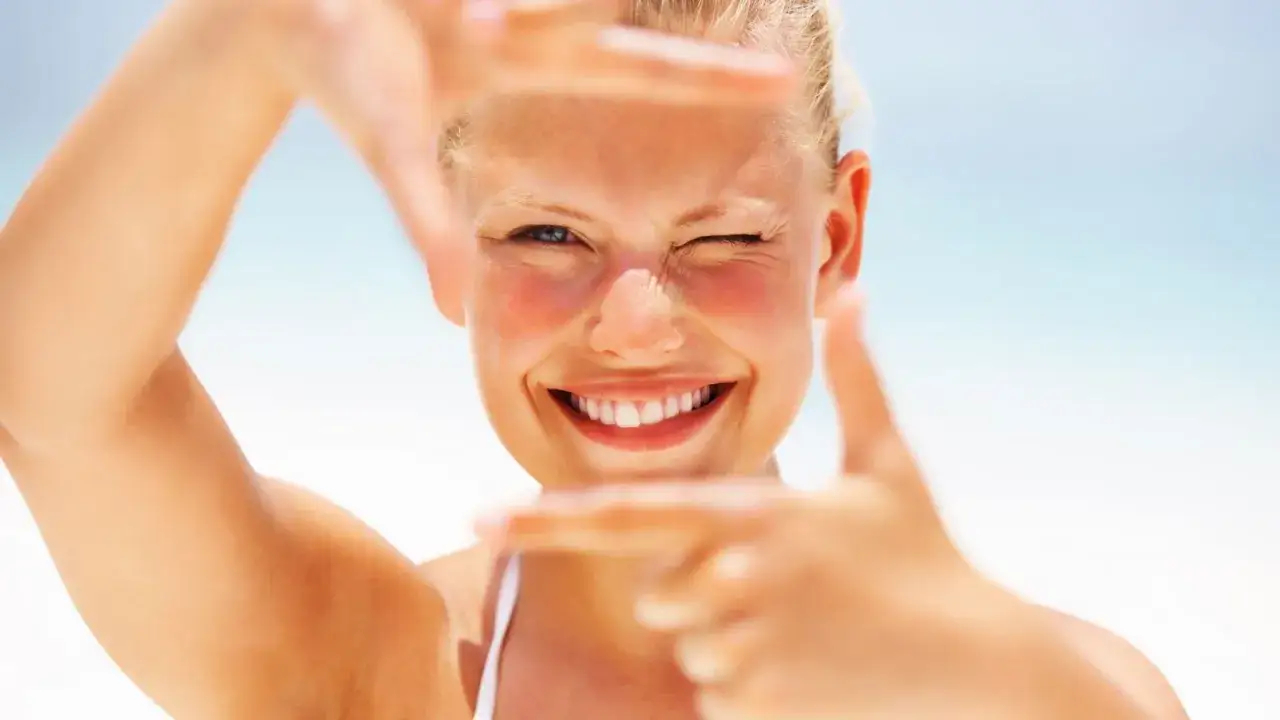 Mujer sonriente guiñando un ojo, con las manos formando un marco. Un ejemplo de cuidados de la piel en verano, con mejillas sonrosadas por el sol.