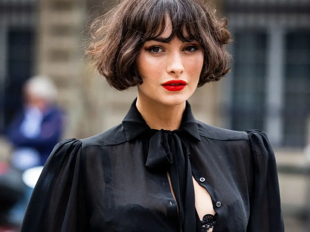 Mujer con un elegante corte bob con flequillo, labios rojos vibrantes y una blusa negra transparente.