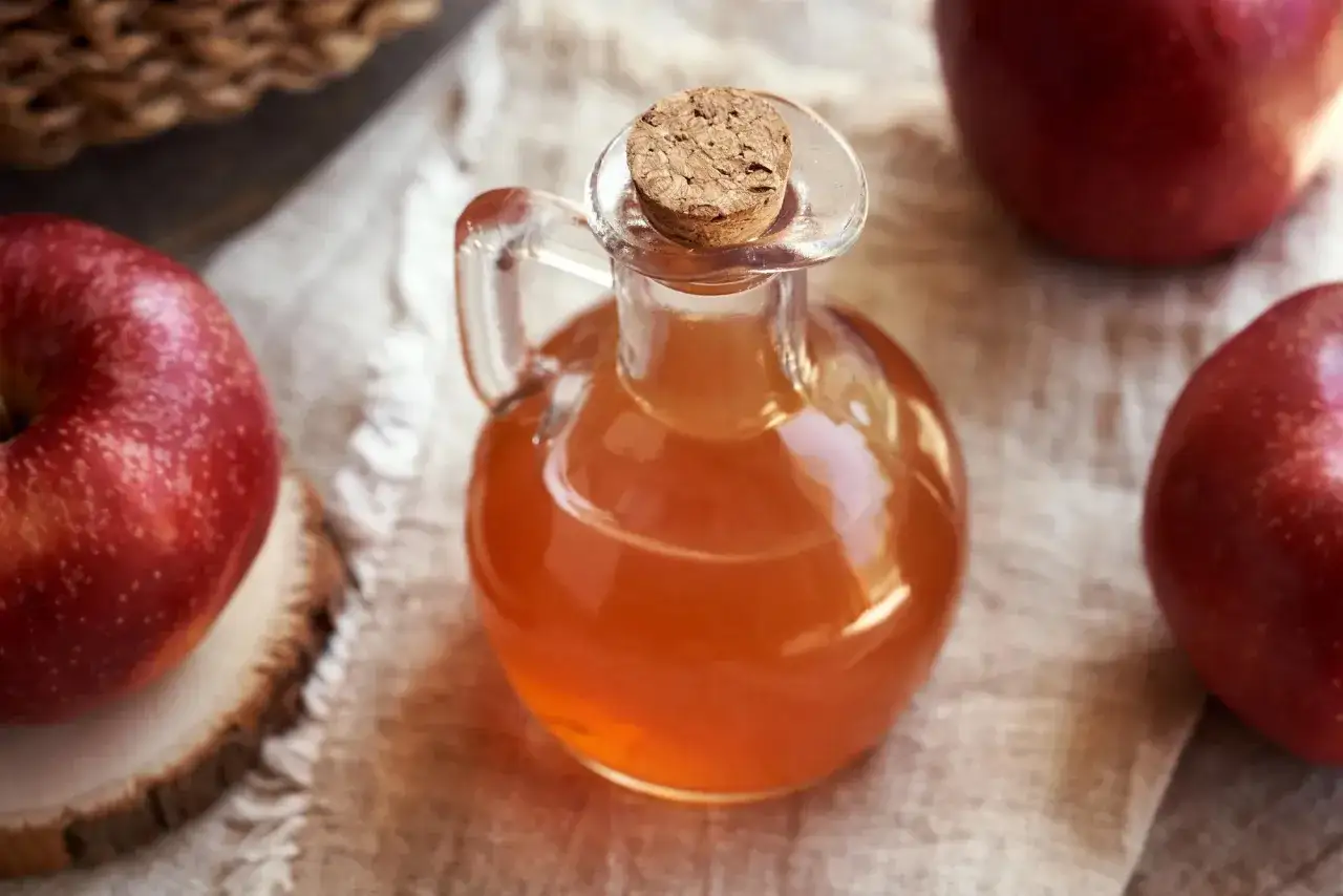 Botella de vidrio con vinagre de manzana para la cara, rodeada de manzanas rojas.