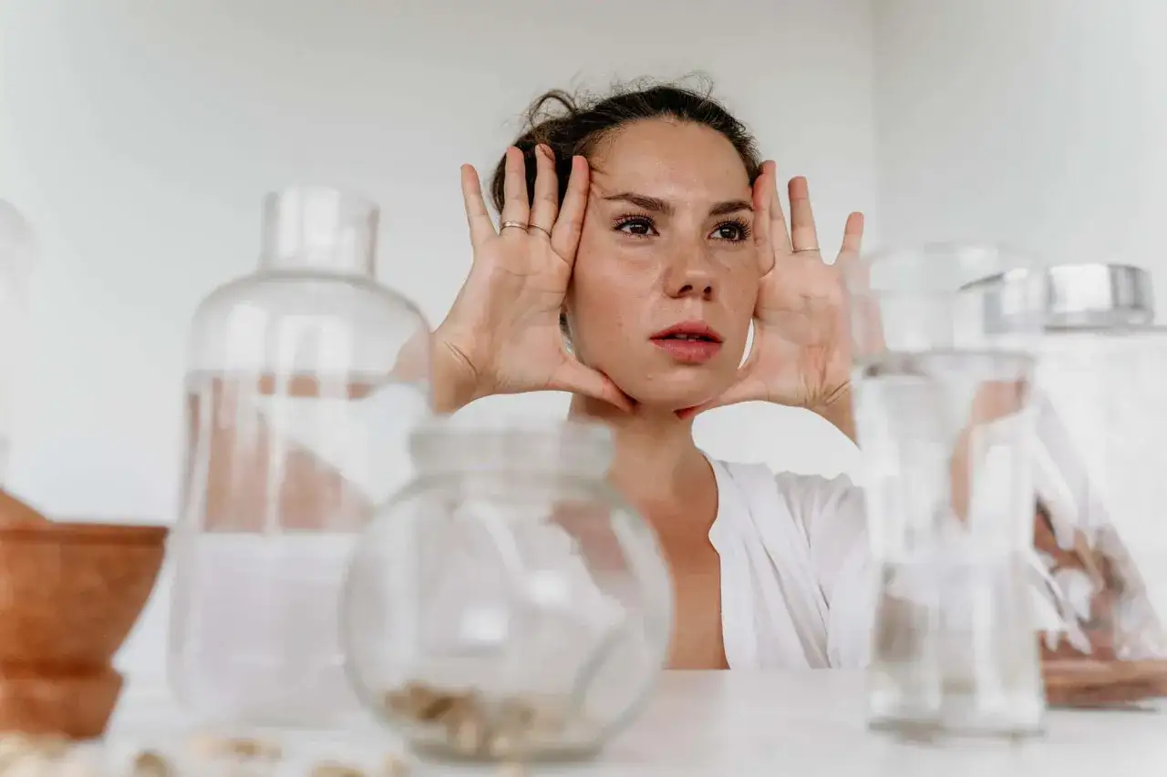 Mujer con manos en el rostro, rodeada de frascos de vidrio, lista para su limpieza facial.