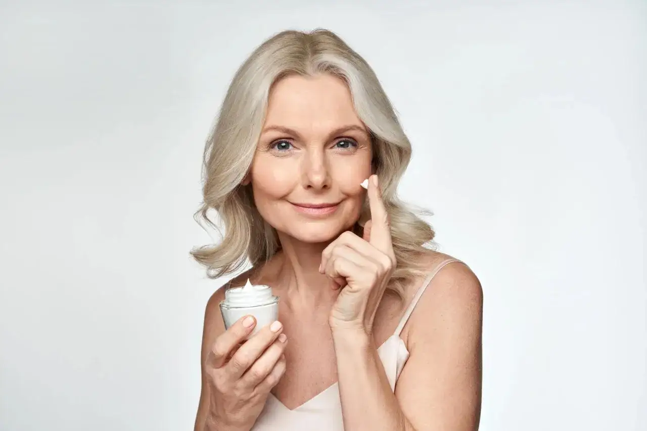 Mujer madura sonr&iacute;e mientras aplica crema, mostrando la belleza del well aging.