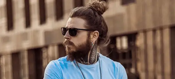 Hombre con pelo largo recogido en un moño, barba, gafas de sol y tatuajes en el cuello.