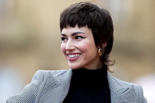Mujer sonríe con un corte pixie medio, luciendo un blazer de pata de gallo y un pendiente dorado.