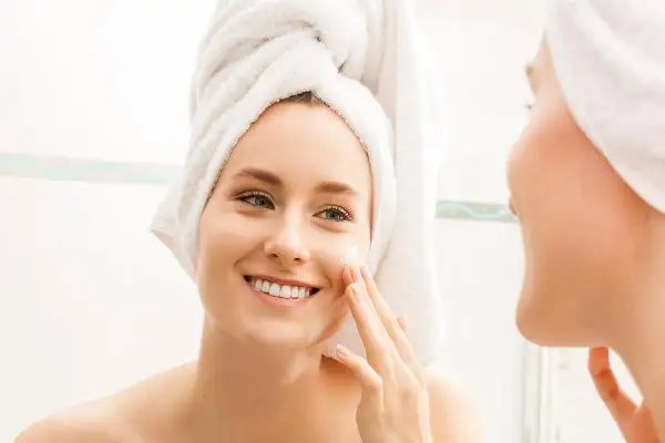 Mujer sonriente con toalla en la cabeza aplicándose crema en la cara. Una rutina de cara para piel grasa que deja la piel radiante.
