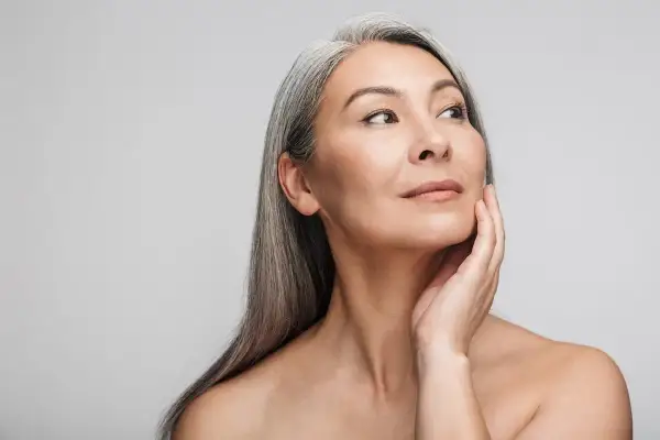 Mujer asiática con cabello gris plateado, tocando su rostro con la mano. Su mirada se dirige hacia la derecha, mostrando una piel radiante y un ejemplo de well aging.