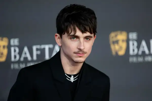 Joven con un corte de pelo hombre medio largo, peinado hacia adelante con flequillo, luciendo un traje negro y un collar brillante.
