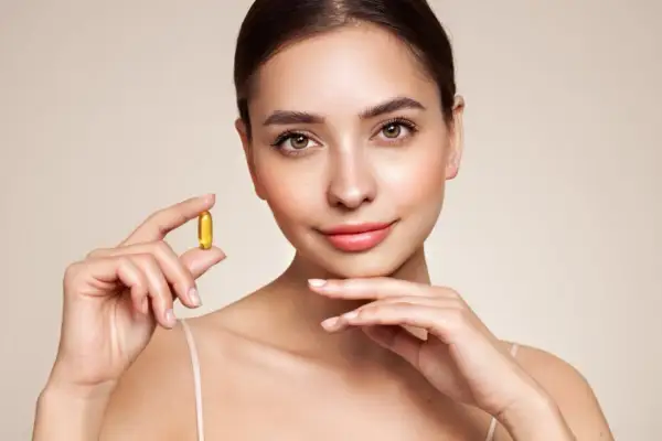 Mujer sonriente sostiene una cápsula dorada, símbolo de vitaminas para la piel, con un fondo neutro.