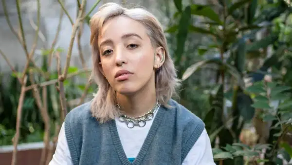 Joven con un moderno corte de pelo mullet mujer, luce un collar plateado y un chaleco azul sobre una camiseta blanca, rodeada de vegetación.