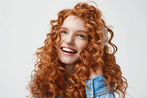 Mujer joven con cabello rojo y rizado, sonriendo y tocándose el pelo. Su melena merodo curly es espectacular.