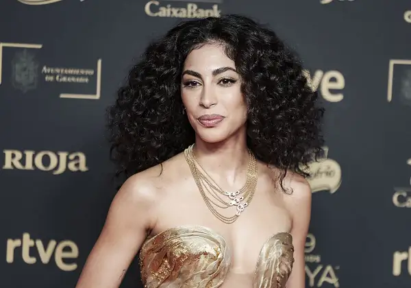 Mujer con cabello oscuro y rizos definidos, luciendo un vestido dorado y un collar de cadenas.
