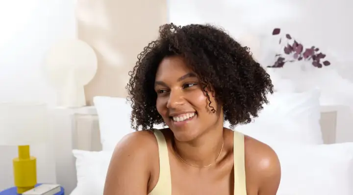 Mujer sonriente con cabello rizado y una permanente ondulada, luciendo un top amarillo en una habitación luminosa.